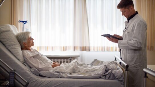 Senior woman lying on hospital bed with her doctor standing by using digital tablet.