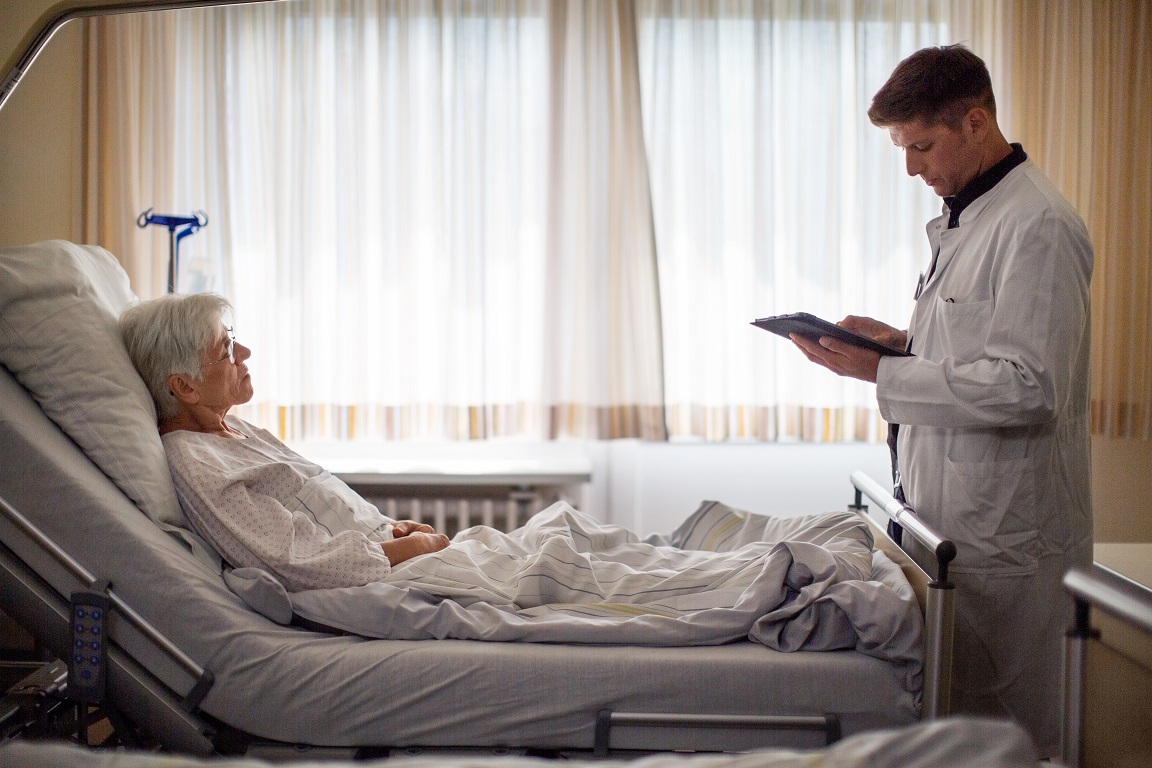 Senior woman lying on hospital bed with her doctor standing by using digital tablet.