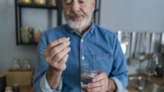 Older man holding glass of water looking at pill in kitchen