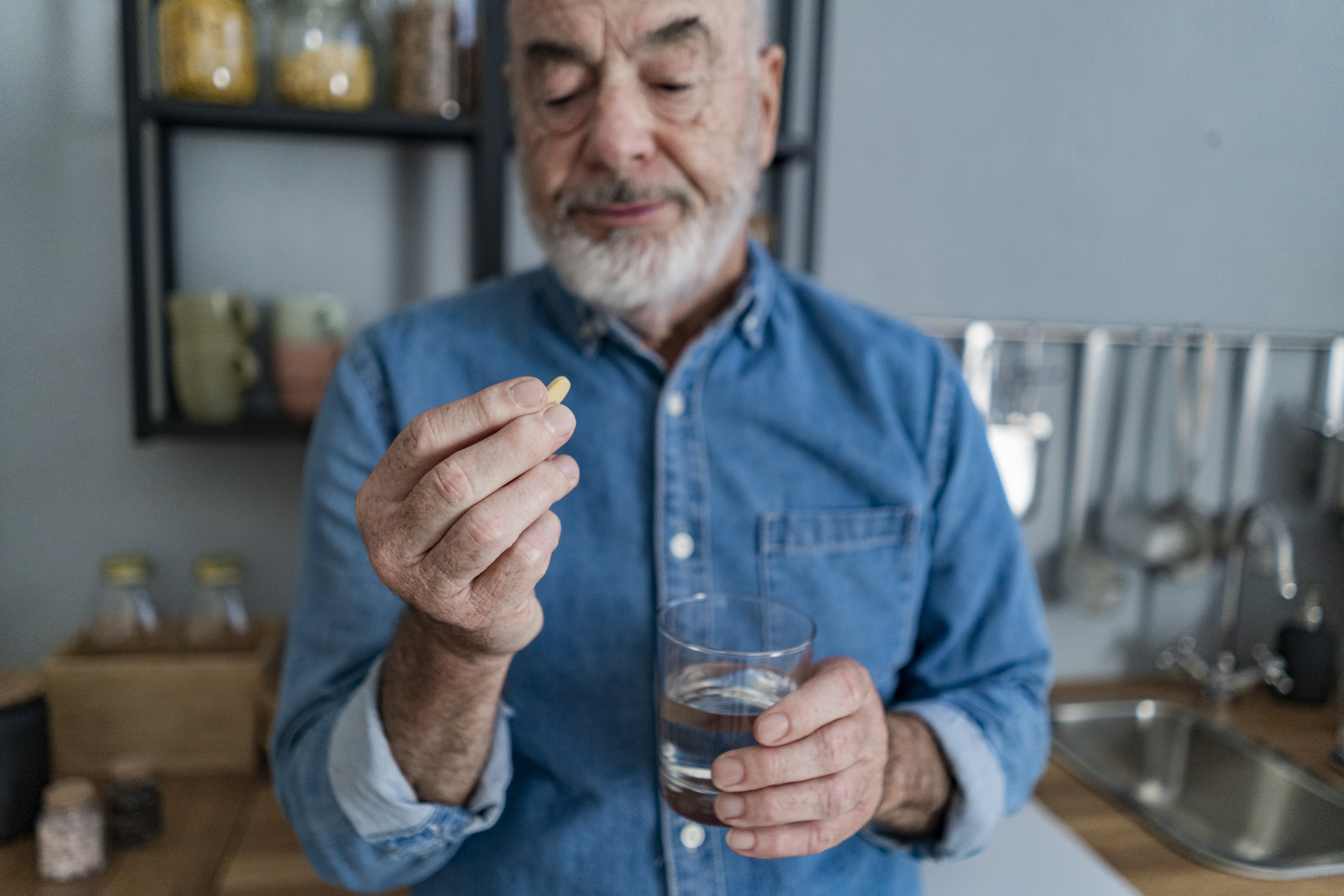 Older man holding glass of water looking at pill in kitchen